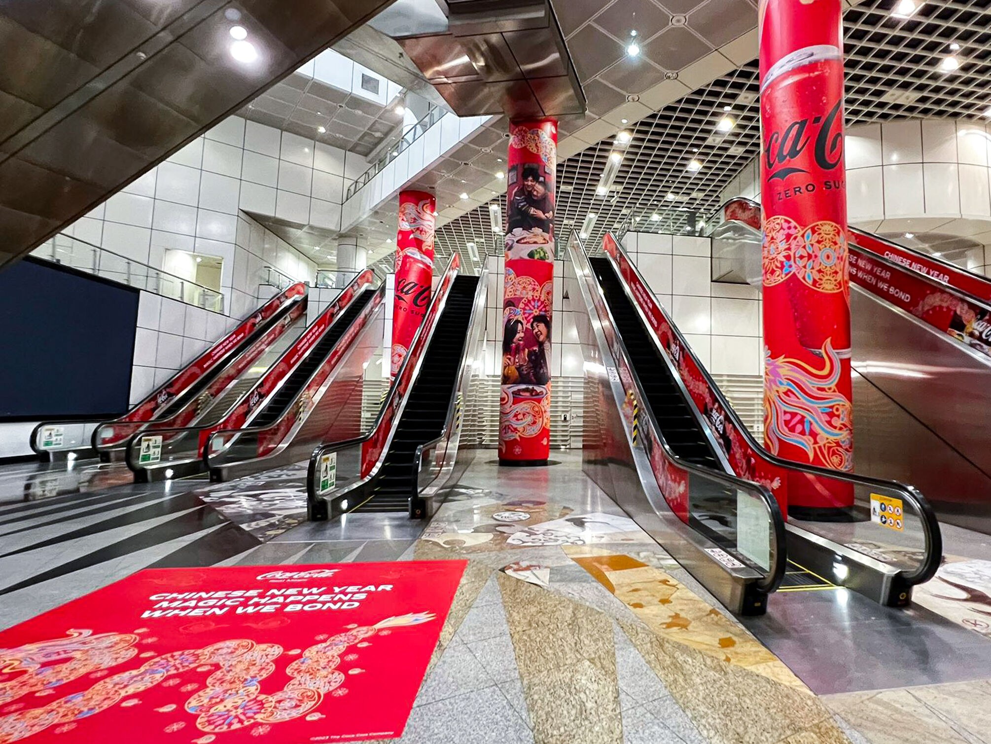 Coca-Cola Chinese New Year mega pillar wraps installed at Dhoby Ghaut MRT, three tall red pillars beside the escalators carrying the dragon motif, a Coca-Cola Zero Sugar can and family dinner portraits, with a matching floor decal in the foreground