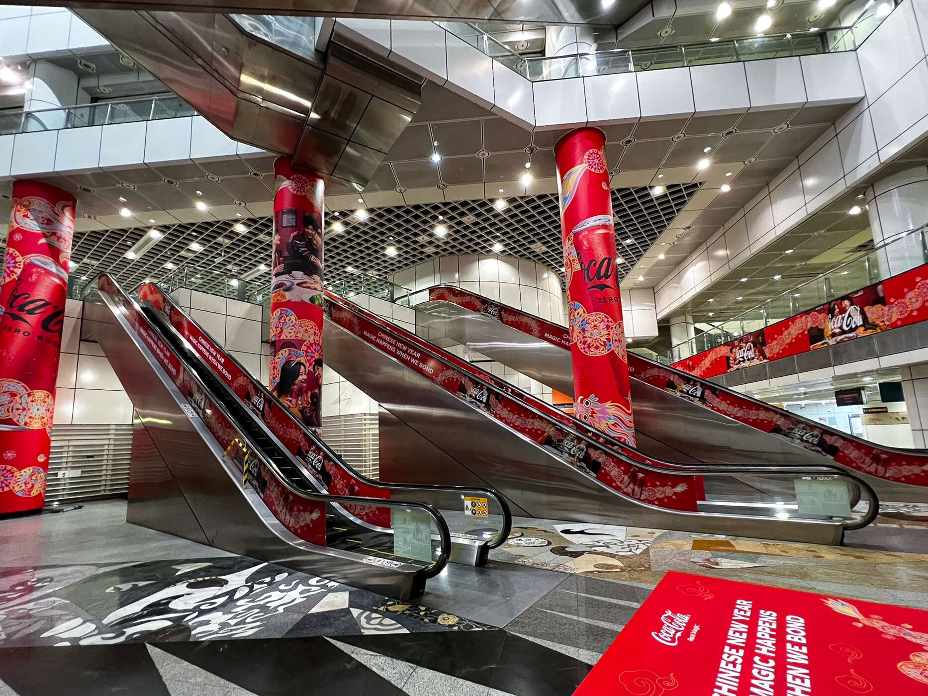 Coca-Cola Chinese New Year mega pillar wraps installed at Dhoby Ghaut MRT, second angle showing the wrapped pillars, escalator side decals and the mezzanine wall graphic